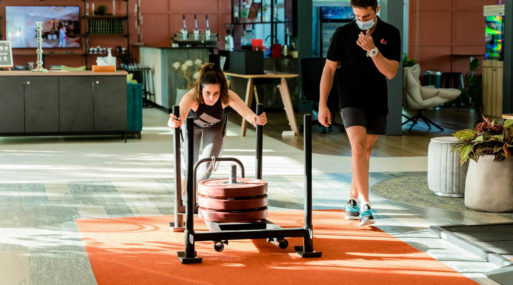 Woman pushing weights on a sled inside a modern gym with trainer observing.