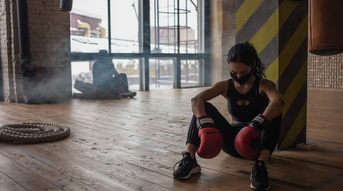 Exhausted African American female fighter in boxing gloves and cloth face mask sitting on parquet against unrecognizable partner in gymnasium